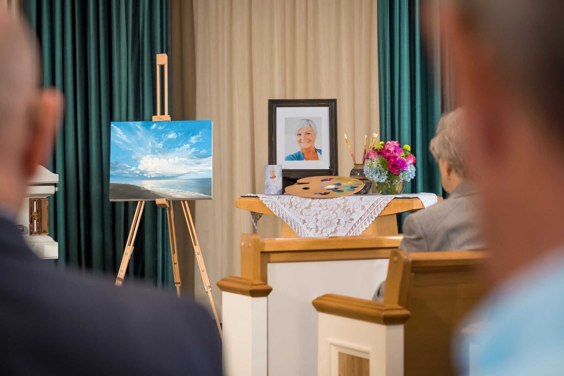 Mourners seated before an easel painting and framed portrait beside flowers in a funeral chapel