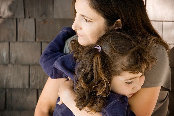 A woman embraces a child with curly hair indoors against a wooden wall.