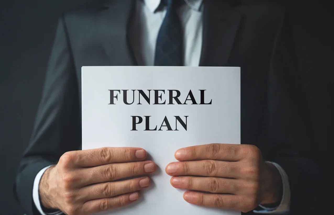 Person in a dark suit holding a paper labeled “FUNERAL PLAN”